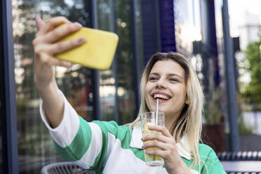 Happy young woman holding drinking glass taking selfie through mobile phone - WPEF06712