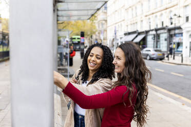 Happy friends touching wall on footpath at bus stop - WPEF06746