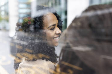 Lächelnde Frau, die mit einem Freund an der Bushaltestelle sitzt, gesehen durch Glas - WPEF06748