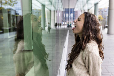 Happy woman sticking out tongue looking at reflection on glass wall - WPEF06767