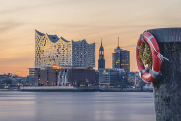 Germany, Hamburg, Life belt hanging on coastal post at dusk with Elbphilharmonie in background - KEBF02480