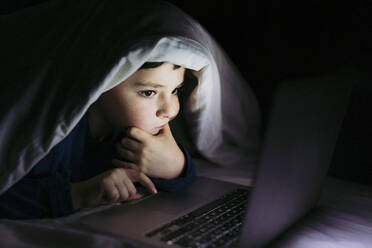 Nachdenklicher Junge schaut auf einen Laptop, der unter einer Decke auf dem Bett liegt - LJF02322