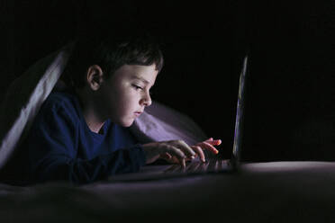 Boy using laptop lying under blanket at home - LJF02324