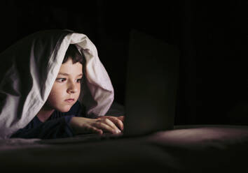 Junge benutzt Laptop unter einer Decke auf dem Bett zu Hause - LJF02325