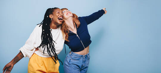 Two happy best friends laughing and having a good time while embracing each other in a studio. Two cheerful female friends making fun memories while standing against a blue background. - JLPSF28303