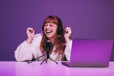 Female podcaster laughing while hosting a live broadcast in a studio. Happy young woman recording an audio show in neon purple light. Woman creating content for her internet podcast. - JLPSF28310