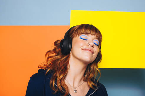Unbeschwerte Frau, die entspannende Musik mit geometrischen Formen im Hintergrund hört. Frau mit rothaarigem Haar und blauem Lidschatten, die eine Audio-Wiedergabeliste über ihre drahtlosen Kopfhörer genießt. - JLPSF28330