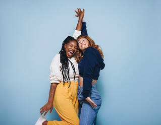 Two best friends dancing and having a good time together in a studio. Happy young women enjoying themselves while standing against a blue background. Female friends making cheerful memories. - JLPSF28333