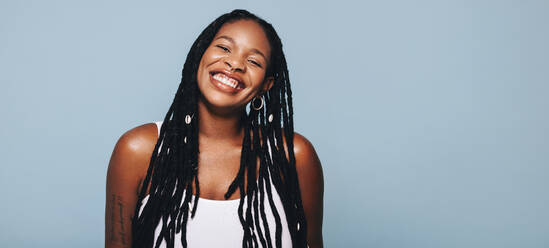 Portrait of a african woman with dreadlocks and body piercings smiling at the camera happily. Cheerful young woman feeling confident in her style. Fashionable woman standing against a studio background. - JLPSF28369