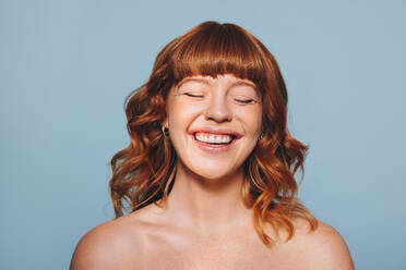 Happy woman with ginger hair and flawless skin smiling with her eyes closed. Cheerful young woman embracing her natural beauty. Body confident young woman standing against a blue studio background. - JLPSF28373