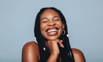 Fröhlich lächelnde Afrikanerin mit Gesichtspiercings in einem Studio. Glückliche junge Frau mit Dreadlocks und Piercing-Schmuck vor blauem Hintergrund. Hipsterin, die sich ihres Stils sicher ist. - JLPSF28380