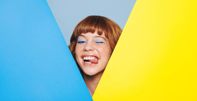 Quirky woman with ginger hair and blue eyeshadow sticking her tongue out while standing behind colour placards. Playful young woman peeping from behind two advertisement boards in a studio. - JLPSF28386
