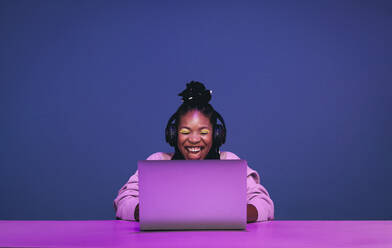Fröhliche weibliche Spielerin, die aufgeregt lächelt, während sie ein Videospiel auf einem Laptop spielt. Glückliche junge Frau, die ein Online-Spiel in einem Studio gewinnt. Junge schwarze Frau, die ein Spiel vor einem lila Hintergrund streamt. - JLPSF28398