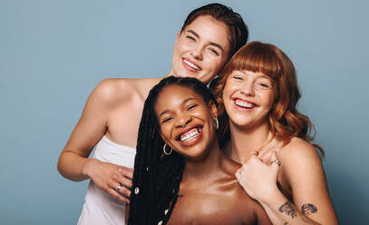 Cheerful women with different skin tones smiling at the camera in a studio. Group of happy young women embracing their natural skin. Portrait of three body positive young women standing together. - JLPSF28409