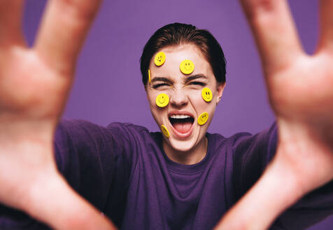 Skurrile junge Frau, die ein Selfie mit Smiley-Aufklebern auf ihrem Gesicht macht. Verspielte Frau, die fröhlich lächelt, während sie ein Foto von sich selbst macht. Glückliche junge Frau, die Spaß vor einem lila Hintergrund hat. - JLPSF28410