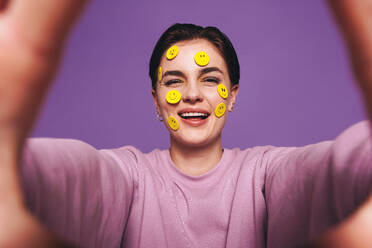 Playful young woman taking a selfie with smiley stickers on her face. Happy woman smiling at the camera while taking a picture of herself. Cheerful young woman having fun against a purple background. - JLPSF28411