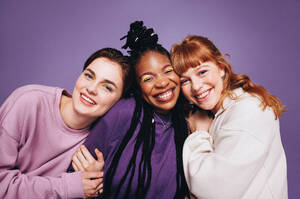 Portrait of happy female friends smiling at the camera in a studio. Best friends enjoying themselves while standing against a purple background. Three young women making happy memories together. - JLPSF28416