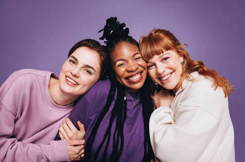 Portrait of happy female friends smiling at the camera in a studio. Best friends enjoying themselves while standing against a purple background. Three young women making happy memories together. - JLPSF28416