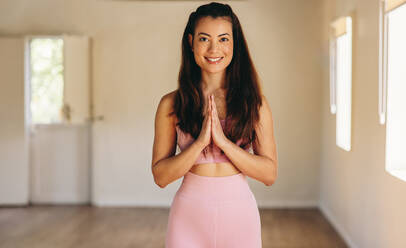 Porträt einer glücklichen jungen Frau, die in Gebetshaltung in die Kamera lächelt. Fröhliche junge Frau, die Hatha-Yoga in einem Yogastudio praktiziert. - JLPSF28429