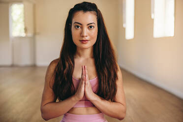 Junge Frau schaut in die Kamera, während sie in einem Yogastudio in Gebetshaltung meditiert. Porträt einer fitten jungen Frau, die eine Atem- und Entspannungsübung praktiziert. - JLPSF28435