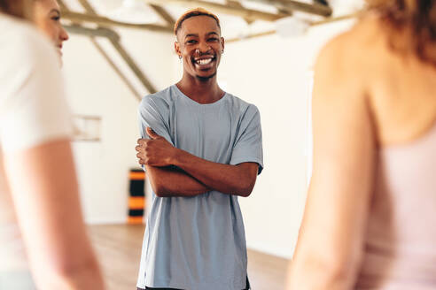 Ein glücklicher junger Mann lächelt in die Kamera, während er mit seiner Yogaklasse im Kreis steht. Eine Gruppe multikultureller Fitness-Freunde, die sich in einem Gemeinschafts-Yogastudio miteinander unterhalten. - JLPSF28449