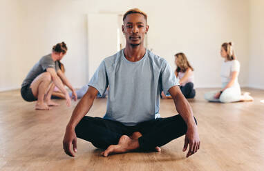 Porträt eines männlichen Yogi, der in die Kamera schaut, während er im Lotussitz sitzt, mit einer Gruppe von Menschen im Hintergrund. Junger schwarzer Mann, der einen Yogakurs in einem Fitnessstudio besucht. - JLPSF28460