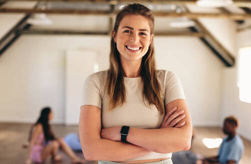 Porträt einer sportlichen jungen Frau, die mit verschränkten Armen in die Kamera lächelt, während sie in einem Fitnesskurs steht. Glückliche junge Frau, die mit ihrer Klasse in einem Yogastudio eine Übungsstunde abhält. - JLPSF28461