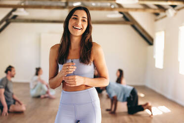 Eine lächelnde Yogalehrerin in einem Yogastudio mit ihrer Klasse im Hintergrund. Eine glückliche junge Frau, die mit einer Gruppe von Menschen in einem Fitnessstudio trainiert. - JLPSF28470