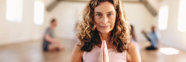 Panoramabild einer reifen Frau, die in Gebetshaltung meditiert, mit einer Gruppe von Menschen im Hintergrund. Ältere Frau, die mit ihrer Klasse in einem Gemeinschafts-Fitnessstudio Yoga praktiziert. - JLPSF28475
