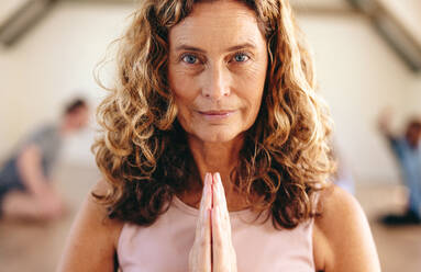 Porträt einer älteren Frau, die in Gebetshaltung meditiert, mit einer Gruppe von Menschen im Hintergrund. Ältere Frau, die Hatha-Yoga mit ihrer Klasse in einem Gemeinschafts-Fitnessstudio praktiziert. - JLPSF28476