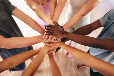 Top view of a group of multicultural people putting their hands together in a huddle before yoga. Group of sporty young people starting a yoga class in a community fitness studio. - JLPSF28499