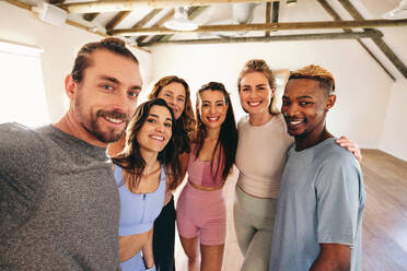 Group of happy fitness friends smiling at the camera while taking a selfie together in a yoga studio. Multicultural sporty people starting a yoga class in a community fitness studio. - JLPSF28507