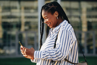 Lächelnde Geschäftsfrau, die auf dem Weg zur Arbeit in der Stadt ein Smartphone benutzt. Seitenansicht einer glücklichen jungen Geschäftsfrau, die morgens auf dem Weg ins Büro eine Textnachricht beantwortet. - JLPSF28518