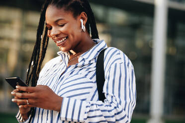 Cheerful young business woman using a smartphone on her way to work in the city. Happy young businesswoman playing music on her phone while commuting to the office in the morning. - JLPSF28519