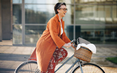 Fröhliche Geschäftsfrau lächelt glücklich, während sie morgens mit dem Fahrrad zur Arbeit fährt. Reife Geschäftsfrau, die ihren umweltfreundlichen Büroweg in der Stadt genießt. - JLPSF28526