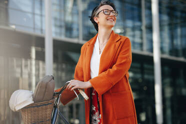 Happy businesswoman looking away thoughtfully while pushing her bike in the city. Smiling mature businesswoman commuting to work with a bicycle in the morning. - JLPSF28528