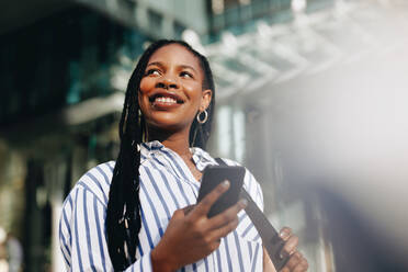 Pensive young business woman looking away with a smile while commuting to work in the city. Happy young business woman using a smartphone on her way to her office in the morning. - JLPSF28535