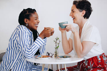 Zwei fröhliche Geschäftsfrauen lachen und trinken gemeinsam Kaffee in einem Bürocafé. Zwei fröhliche Geschäftsfrauen genießen ein freundliches Kaffeetreffen an einem modernen Arbeitsplatz. - JLPSF28541