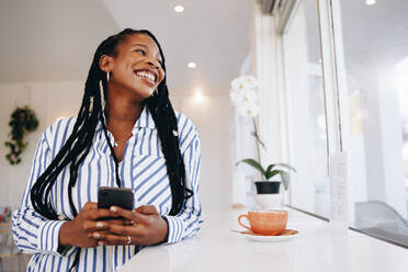 Junge Geschäftsfrau lächelt und schaut weg, während sie ein Smartphone in einem Café hält. Glückliche junge schwarze Geschäftsfrau, die ihre Kaffeepause in einem Café genießt. - JLPSF28544