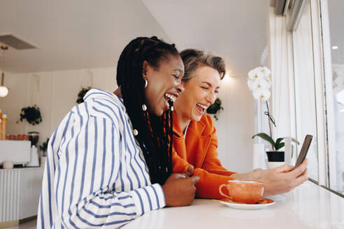 Zwei glückliche Geschäftsfrauen, die in einem Café per Smartphone Videotelefonate mit ihren Kollegen führen. Fröhliche Geschäftsfrauen, die sich morgens virtuell mit ihrem Team treffen, während sie aus der Ferne arbeiten. - JLPSF28547