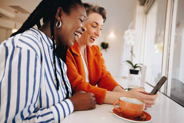 Zwei glückliche Geschäftsfrauen, die in einem Café per Smartphone Videotelefonate mit ihrem Team führen. Lächelnde Geschäftsfrauen, die sich morgens virtuell mit ihren Kollegen unterhalten, während sie aus der Ferne arbeiten. - JLPSF28550