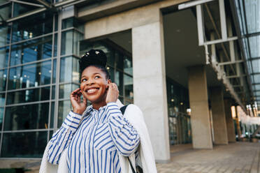 Smiling businesswoman listening to music on earphones during her morning commute in the city. Happy young business woman enjoying a playlist while walking to the office. - JLPSF28553