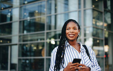 Pensive businesswoman holding a smartphone and wearing earphones during her work commute in the city. Young black business woman listening to music on her way to the office in the morning. - JLPSF28557