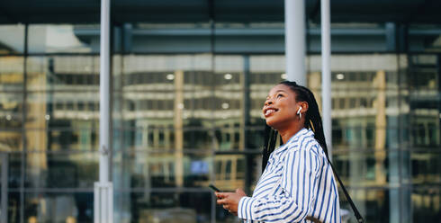 Sideview of a young businesswoman looking away and holding a smartphone while commuting in the city. Cheerful young business woman listening to music on her way to the office in the morning. - JLPSF28558