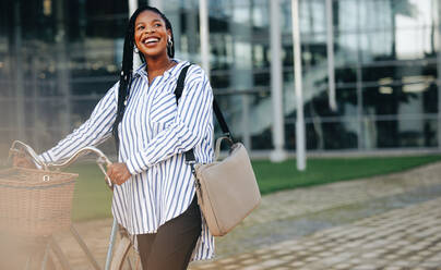 Young businesswoman smiling cheerfully while walking alongside her bicycle in the city. Happy young business woman commuting to the office in the morning. - JLPSF28563