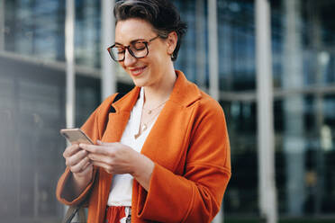 Glückliche Geschäftsfrau, die auf dem Weg zur Arbeit in der Stadt auf ihr Smartphone schaut. Reife Geschäftsfrau, die morgens auf dem Weg ins Büro eine Textnachricht liest. - JLPSF28570