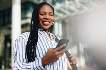 Young business woman smiling at the camera while commuting to her office in the city. Portrait of a happy young business woman using a smartphone on her way to work in the morning. - JLPSF28575