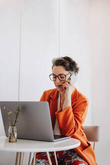 Eine Geschäftsfrau telefoniert, während sie in einem Café an einem Laptop arbeitet. Eine reife Geschäftsfrau macht Pläne mit einem ihrer Kunden, während sie aus der Ferne arbeitet. - JLPSF28583