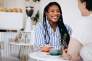 Happy young businesswoman smiling while having a coffee meeting with a colleague in a cafe. Two cheerful businesswomen having a friendly conversation in a modern coffee shop. - JLPSF28588