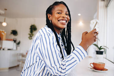Happy young businesswoman smiling at the camera while holding a smartphone in a coffee shop. Cheerful young black businesswoman taking a coffee break in a cafe. - JLPSF28594